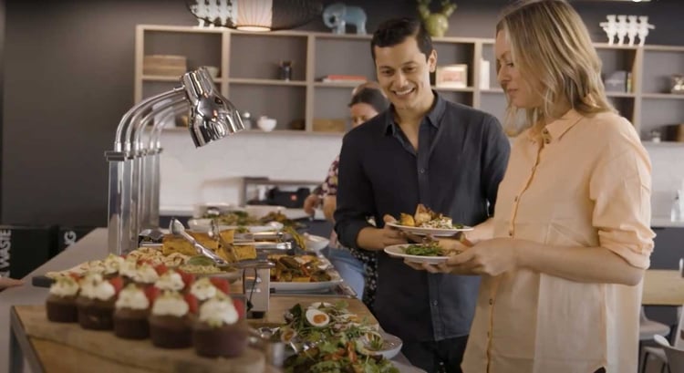 The image shows a workplace with a lunch display and two employees chatting over lunch